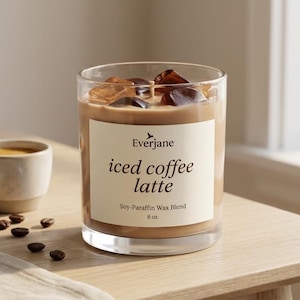 May include: A clear glass candle with iced coffee latte scent, filled with a light brown wax and ice cubes. The candle has a label that reads "iced coffee latte" and is 8 oz. A small cup of coffee and coffee beans are next to the candle.