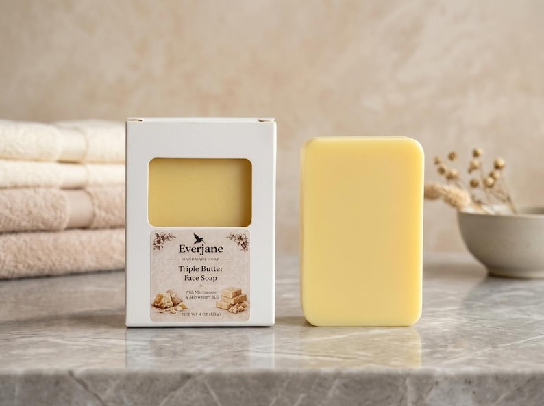 May include: A yellow soap bar and a white box with a label reading "Triple Butter Face Soap" are on a marble surface. The soap is next to the box, which has a window showing the soap inside. The soap weighs 113 grams.