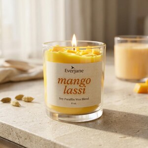 May include: A lit mango lassi scented candle in a clear glass jar. The candle has a yellow wax with mango chunks on top. The label reads "mango lassi" and "Soy-Paraffin Wax Blend 8 oz.". A glass of mango lassi and mango pieces are in the background.