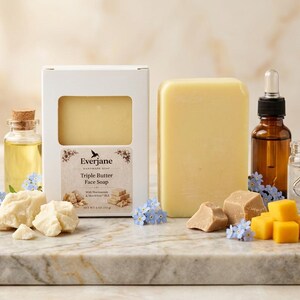 May include: A display of skincare items, including a yellow soap bar, a bottle of oil, and a box of soap labelled "Triple Butter Face Soap." Also shown are butter chunks and cubes, and small blue flowers. The items are arranged on a marble surface.