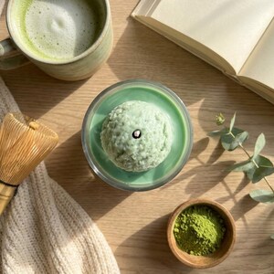 May include: Overhead shot of a matcha-themed setup. A green matcha latte in a ceramic mug, a matcha candle, a bamboo whisk, and a small bowl of matcha powder are arranged on a wooden surface. An open book and a sprig of eucalyptus are also present.