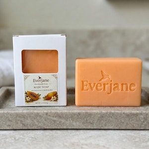 May include: A rectangular orange bar of Everjane handmade Kojic soap with the brand name embossed on the front, accompanied by its white cardboard packaging featuring a cutout window, papaya illustrations, and the text Net Wt 4 oz (113 g).
