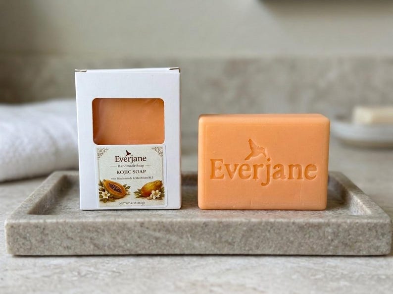 May include: A rectangular orange bar of Everjane handmade Kojic soap with the brand name embossed on the front, accompanied by its white cardboard packaging featuring a cutout window, papaya illustrations, and the text Net Wt 4 oz (113 g).