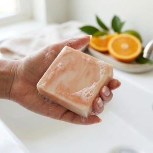 Puede incluir: Una pastilla de jab&oacute;n cuadrada con una paleta de colores jaspeados en tono melocot&oacute;n y blanco, que presenta una ligera capa de espuma transl&uacute;cida en su superficie. El jab&oacute;n tiene una textura suave y s&oacute;lida, sostenido en la palma de la mano.