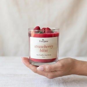 May include: A clear glass candle holder with red wax and fresh strawberries on top. The candle has a label that reads "strawberry bliss" and is a soy-paraffin wax blend, 8 oz.