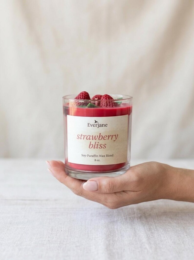 May include: A clear glass candle holder with red wax and fresh strawberries on top. The candle has a label that reads "strawberry bliss" and is a soy-paraffin wax blend, 8 oz.