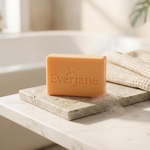 May include: A rectangular peach-colored bar of soap with the word Everjane and a small hummingbird icon embossed on the front. The soap rests on a flat stone surface next to a folded beige waffle-textured towel with visible soap suds.
