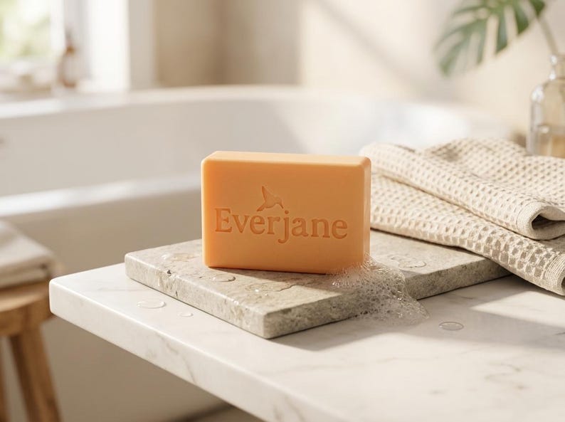 May include: A rectangular peach-colored bar of soap with the word Everjane and a small hummingbird icon embossed on the front. The soap rests on a flat stone surface next to a folded beige waffle-textured towel with visible soap suds.