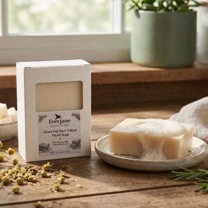Op de afbeelding: Een witte rechthoekige doos Everjane Grass-Fed Beef Tallow gezichtszeep naast een beige stuk zeep met wit schuim op een klein bordje. Het etiket bevat een vogelicoon en de tekst Net Wt 113g.