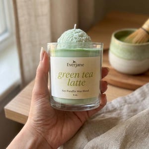 May include: A green tea latte candle in a clear glass, with a soy-paraffin wax blend. The candle has a light green color and a textured top. The label reads "green tea latte" and "8 oz."