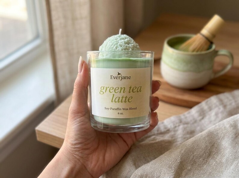 May include: A green tea latte candle in a clear glass, with a soy-paraffin wax blend. The candle has a light green color and a textured top. The label reads "green tea latte" and "8 oz."