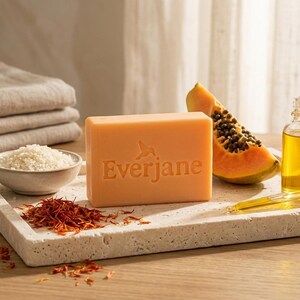 May include: A rectangular peach-colored bar of soap embossed with the text Everjane and a hummingbird icon, accompanied by a slice of papaya, a small bowl of white rice, loose saffron threads, and a glass dropper bottle filled with golden oil.