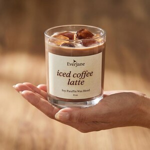 May include: A clear glass candle with a coffee-colored wax, ice cubes, and a label that reads "iced coffee latte." The candle is 8 oz and is a soy-paraffin wax blend. The candle is held in a hand.