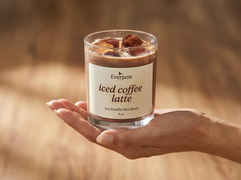 May include: A clear glass candle with a coffee-colored wax, ice cubes, and a label that reads "iced coffee latte." The candle is 8 oz and is a soy-paraffin wax blend. The candle is held in a hand.