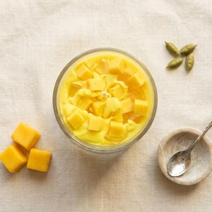May include: A clear glass bowl filled with yellow liquid and topped with yellow cubes. A small wooden bowl with a spoon, and a few green pods are next to the bowl. Three yellow cubes are on the left side of the image.