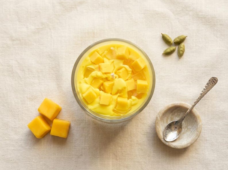May include: A clear glass bowl filled with yellow liquid and topped with yellow cubes. A small wooden bowl with a spoon, and a few green pods are next to the bowl. Three yellow cubes are on the left side of the image.