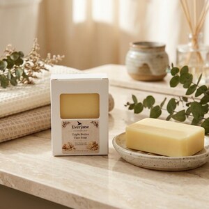 May include: A bar of yellow soap on a speckled ceramic soap dish, beside a white box of soap. The box is labelled "Triple Butter Face Soap". A stack of folded beige towels and decorative greenery are also present.