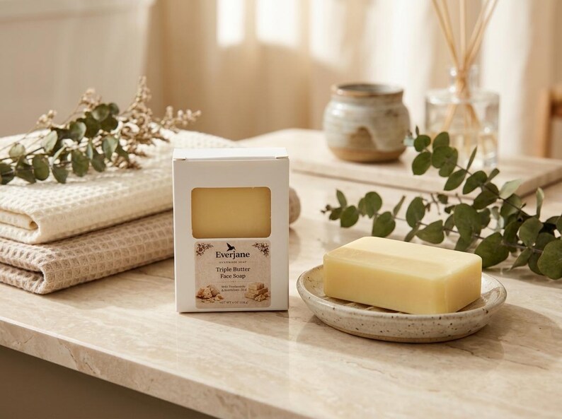 May include: A bar of yellow soap on a speckled ceramic soap dish, beside a white box of soap. The box is labelled "Triple Butter Face Soap". A stack of folded beige towels and decorative greenery are also present.
