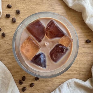 May include: Overhead view of a clear glass candle filled with a creamy, light brown wax. The candle features several large, amber-colored wax cubes resembling ice. Scattered coffee beans and a beige cloth add to the coffee theme.
