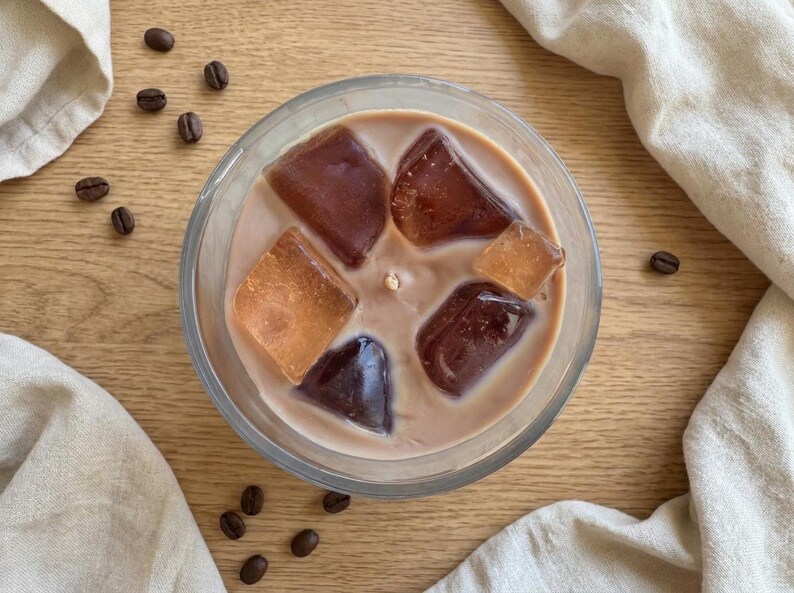 May include: Overhead view of a clear glass candle filled with a creamy, light brown wax. The candle features several large, amber-colored wax cubes resembling ice. Scattered coffee beans and a beige cloth add to the coffee theme.