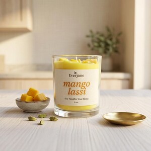 May include: A clear glass candle with yellow wax and the words "mango lassi" on the label. The candle sits on a light wood surface with a small bowl of mango cubes, a gold dish, and cardamom pods.