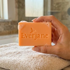 May include: A rectangular bar of peach-colored soap with the word Everjane and a hummingbird icon embossed on the front. The soap surface has small bubbles and moisture, indicating it is wet and ready for use in a bathroom setting.