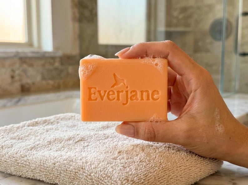 May include: A rectangular bar of peach-colored soap with the word Everjane and a hummingbird icon embossed on the front. The soap surface has small bubbles and moisture, indicating it is wet and ready for use in a bathroom setting.