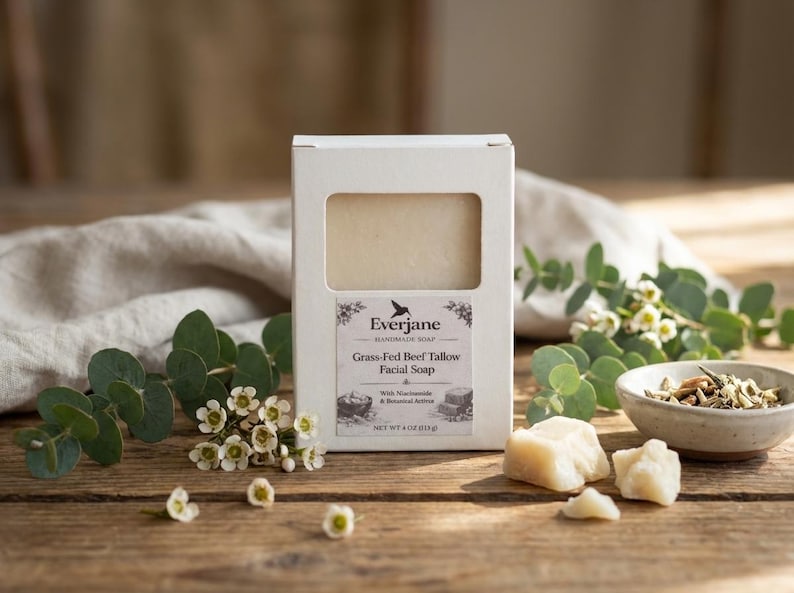 Op de afbeelding: Een rechthoekige witte doos met een handgemaakt Everjane gezichtszeepblok. Het etiket vermeldt Grass-Fed Beef Tallow Facial Soap with Niacinamide & Botanical Actives, Net Wt 4 oz (113 g). De zeep is lichtbeige van kleur en solide.