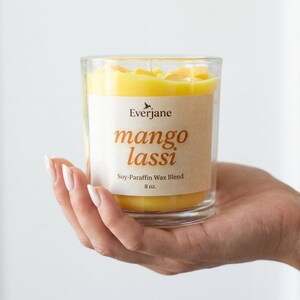 May include: A clear glass candle holder filled with yellow wax. The label reads "Everjane mango lassi Soy-Paraffin Wax Blend 8 oz."