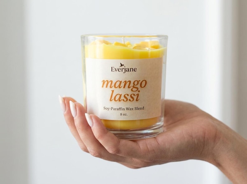 May include: A clear glass candle holder filled with yellow wax. The label reads "Everjane mango lassi Soy-Paraffin Wax Blend 8 oz."