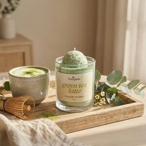 May include: A green tea latte candle with a matcha whisk and a cup of matcha latte on a wooden tray. The candle has a green wax blend and the text "green tea latte" on the label. The candle is 4 oz.
