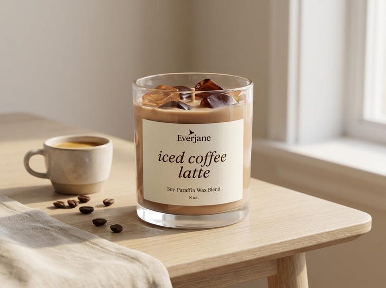 May include: A clear glass candle with iced coffee latte scent, filled with a light brown wax and ice cubes. The candle has a label that reads "iced coffee latte" and is 8 oz. A small cup of coffee and coffee beans are next to the candle.