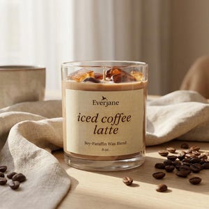 May include: A clear glass candle with a light brown wax, labeled "iced coffee latte." The candle has ice cubes on top and is surrounded by coffee beans. A beige mug and a linen cloth are in the background. The candle is 8 oz.