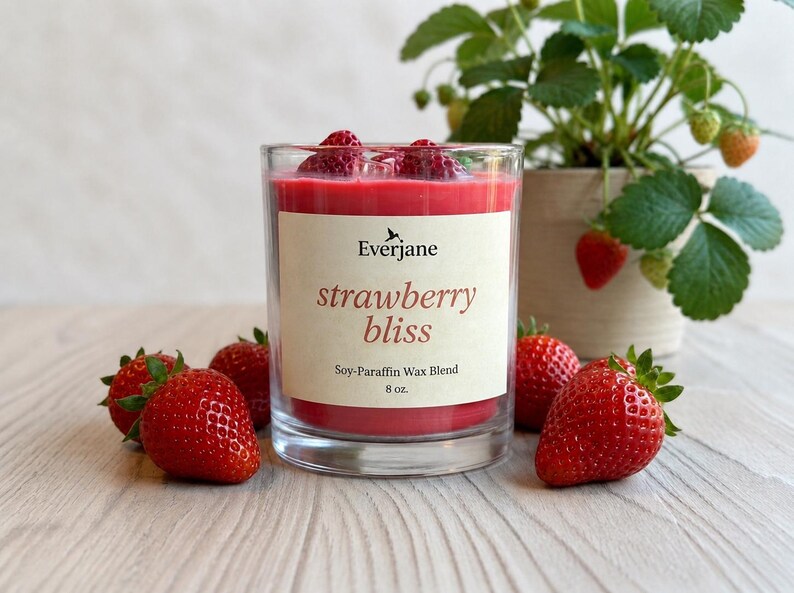 May include: A clear glass candle with red wax and fresh strawberries on top, labeled "strawberry bliss." The candle is surrounded by fresh strawberries and a potted strawberry plant. The candle is 8 oz.