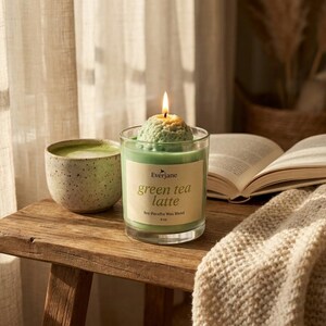May include: A lit green tea latte scented candle in a glass jar, next to a ceramic cup of matcha and an open book. A cozy beige blanket is draped over the wooden table. The candle is 3 inches tall.