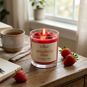 May include: A lit strawberry-scented candle in a clear glass jar with the words "strawberry bliss" on the label. Fresh strawberries and a mug of tea sit on a wooden table next to an open notebook and pen.