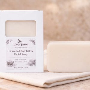 Op de afbeelding: Een rechthoekig stuk gebroken witte gezichtszeep naast de witte kartonnen verpakking. Het etiket vermeldt Everjane Handmade Soap, Grass-Fed Beef Tallow Facial Soap, With Niacinamide & Botanical Actives en Net Wt 4 oz (115g).