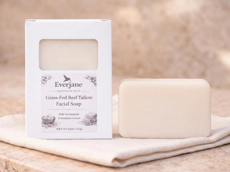 Op de afbeelding: Een rechthoekig stuk gebroken witte gezichtszeep naast de witte kartonnen verpakking. Het etiket vermeldt Everjane Handmade Soap, Grass-Fed Beef Tallow Facial Soap, With Niacinamide & Botanical Actives en Net Wt 4 oz (115g).