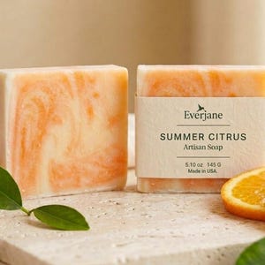 Puede incluir: Dos pastillas de jab&oacute;n rectangulares con un patr&oacute;n jaspeado en blanco y naranja. Una pastilla tiene una etiqueta de papel que dice Everjane SUMMER CITRUS Artisan Soap 5.10 oz 145 G Made in USA. El jab&oacute;n se muestra con rodajas de naranja y hojas verdes.