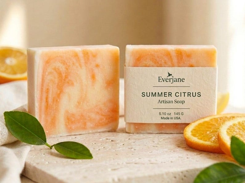 Puede incluir: Dos pastillas de jab&oacute;n rectangulares con un patr&oacute;n jaspeado en blanco y naranja. Una pastilla tiene una etiqueta de papel que dice Everjane SUMMER CITRUS Artisan Soap 5.10 oz 145 G Made in USA. El jab&oacute;n se muestra con rodajas de naranja y hojas verdes.