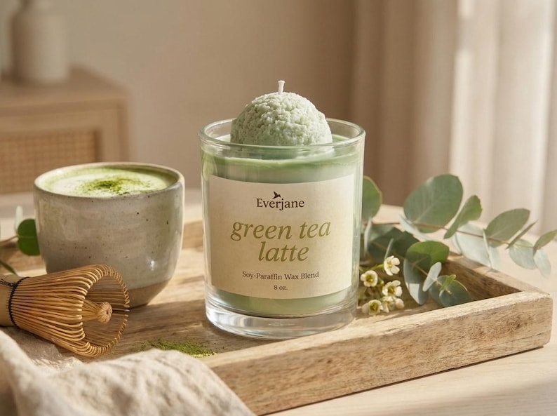 May include: A green tea latte candle with a soy-paraffin wax blend, 8 oz. The candle has a green, spherical top and is in a clear glass. A ceramic mug with matcha and a bamboo whisk are also in the image.