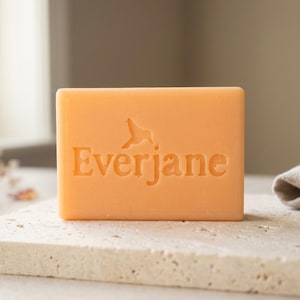 May include: A rectangular bar of peach-colored soap featuring the embossed text Everjane with a small bird icon above the letters. The soap has a smooth, solid surface and is positioned upright, displaying the brand name clearly on its front face.