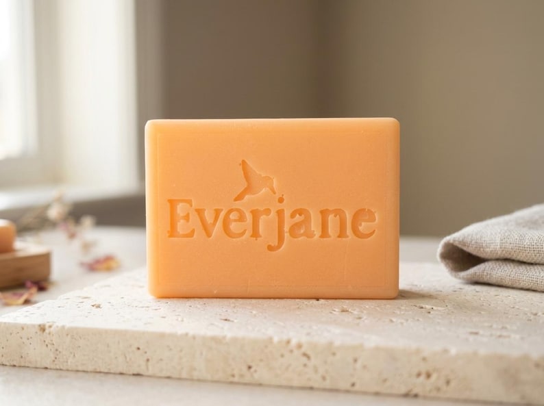 May include: A rectangular bar of peach-colored soap featuring the embossed text Everjane with a small bird icon above the letters. The soap has a smooth, solid surface and is positioned upright, displaying the brand name clearly on its front face.
