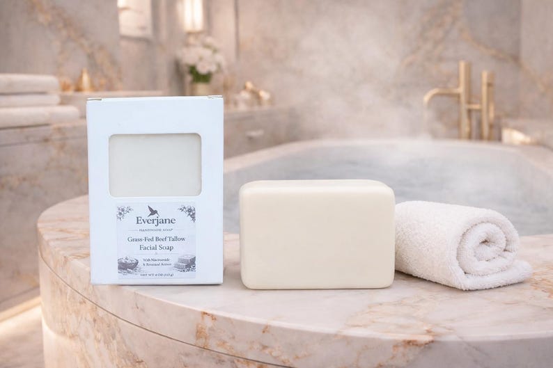 Op de afbeelding: Een rechthoekig blok gebroken witte gezichtszeep naast de witte kartonnen verpakking. De doos heeft een uitgesneden venster, de merknaam Everjane en de tekst Grass-Fed Beef Tallow Facial Soap. Een kleine opgerolde witte handdoek ligt naast de zeep.