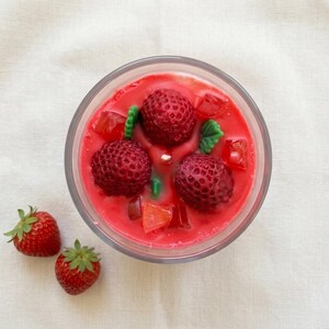 May include: A clear glass candle filled with red wax, featuring three large, realistic strawberry-shaped wax pieces and smaller wax cubes. Two fresh strawberries with green stems sit beside the candle on a white surface.