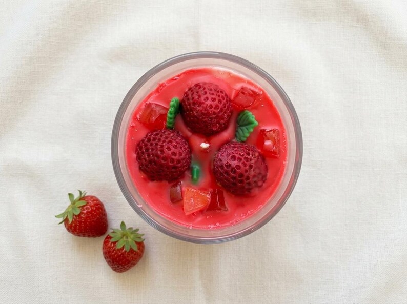 May include: A clear glass candle filled with red wax, featuring three large, realistic strawberry-shaped wax pieces and smaller wax cubes. Two fresh strawberries with green stems sit beside the candle on a white surface.