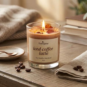 May include: A lit candle in a glass jar with "iced coffee latte" label. The candle has a coffee-colored wax and is surrounded by coffee beans, a small plate with a spoon, and a folded cloth. The candle is 8 oz.