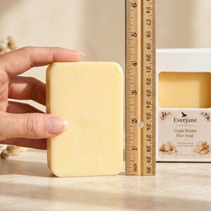 May include: A rectangular, light yellow face soap, approximately 7.6 cm in length, is held next to a ruler. The soap is in a white box with the brand name "Everjane" and "Triple Butter Face Soap" printed on it. A natural sponge is in the background.