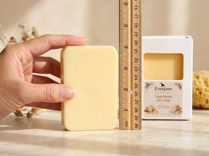 May include: A rectangular, light yellow face soap, approximately 7.6 cm in length, is held next to a ruler. The soap is in a white box with the brand name "Everjane" and "Triple Butter Face Soap" printed on it. A natural sponge is in the background.