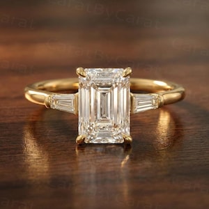 May include: A gold ring with a large, rectangular, clear gemstone as the centerpiece. Two smaller, rectangular gemstones flank the main stone. The ring is set on a dark wooden surface.